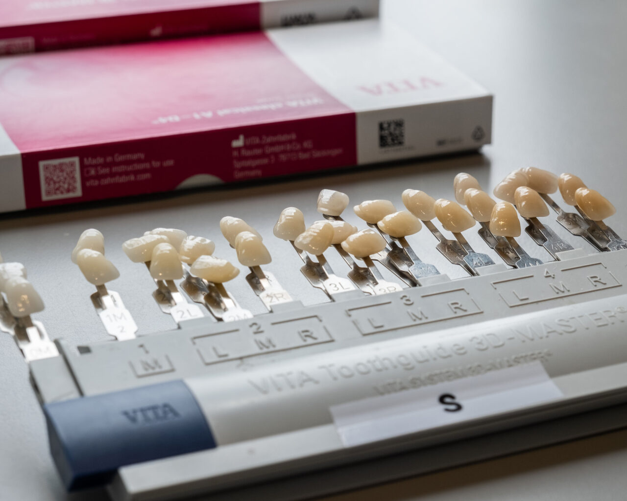 A dental shade guide displays various tooth-colored samples labeled with codes, used to match the color of dental restorations. Two pink and white VITA product boxes are in the background.