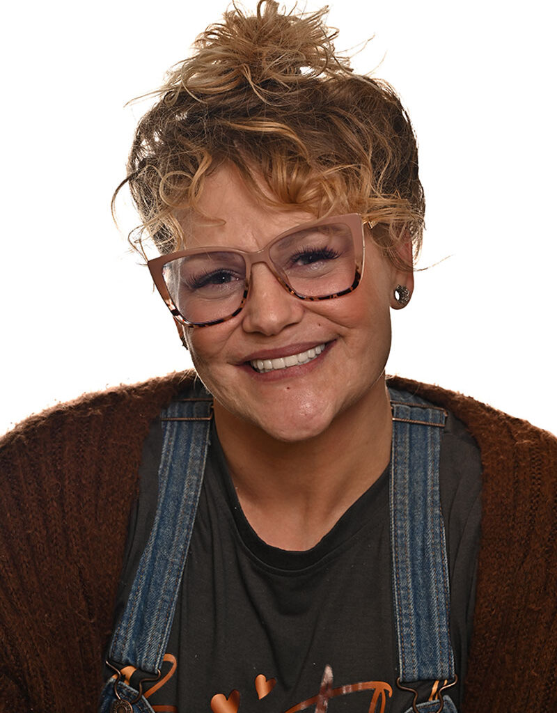 'http://A%20smiling%20person%20with%20curly%20blonde%20hair%20in%20a%20messy%20bun,%20wearing%20oversized%20clear-framed%20glasses,%20denim%20overalls,%20a%20brown%20sweater,%20and%20a%20black%20t-shirt,%20posing%20against%20a%20white%20background.