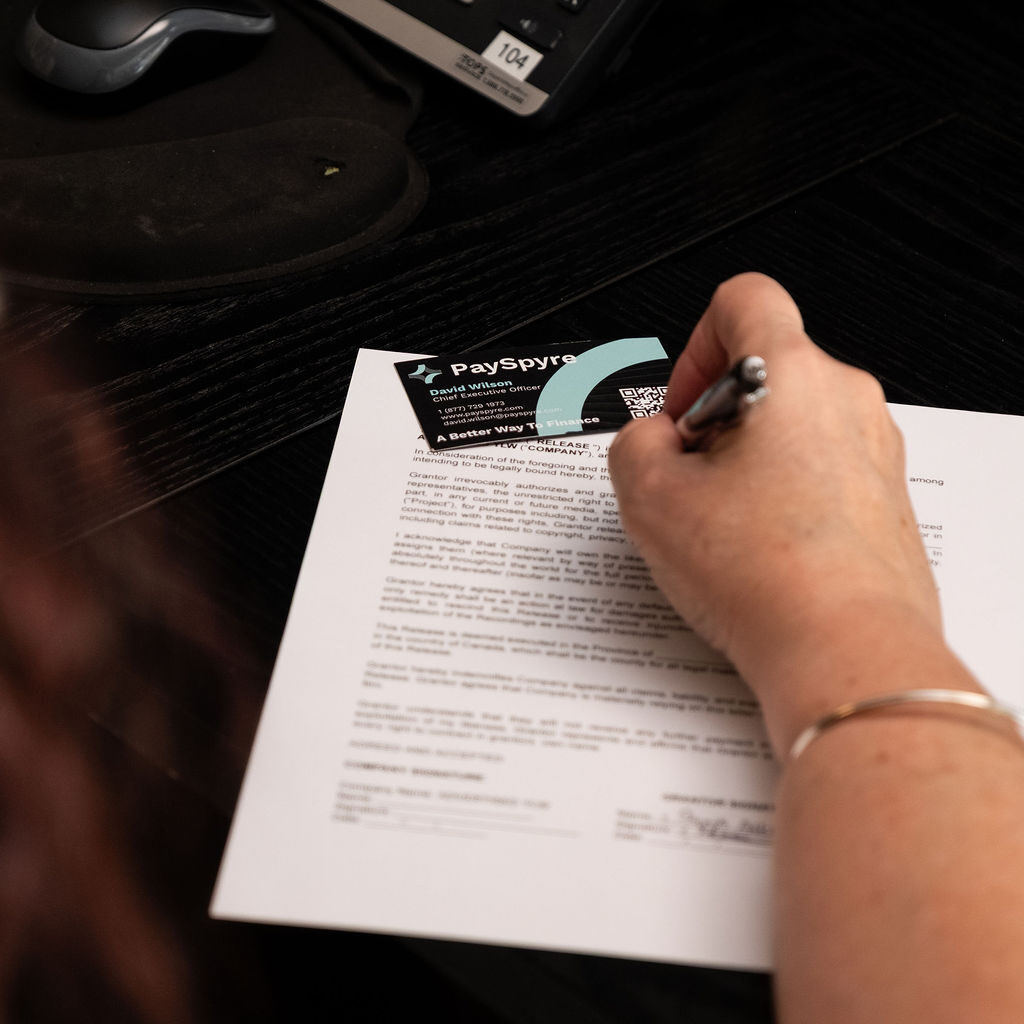 'http://A%20person%20is%20signing%20a%20printed%20document%20labeled%20PaySpyre%20on%20a%20dark%20desk,%20with%20part%20of%20a%20keyboard%20and%20a%20mouse%20pad%20visible%20in%20the%20background.
