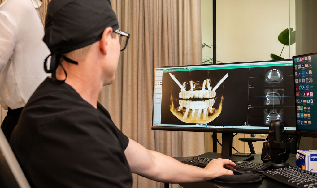 A person in medical scrubs and a cap views dental X-ray images of teeth and jaw implants on a computer monitor in an office setting.