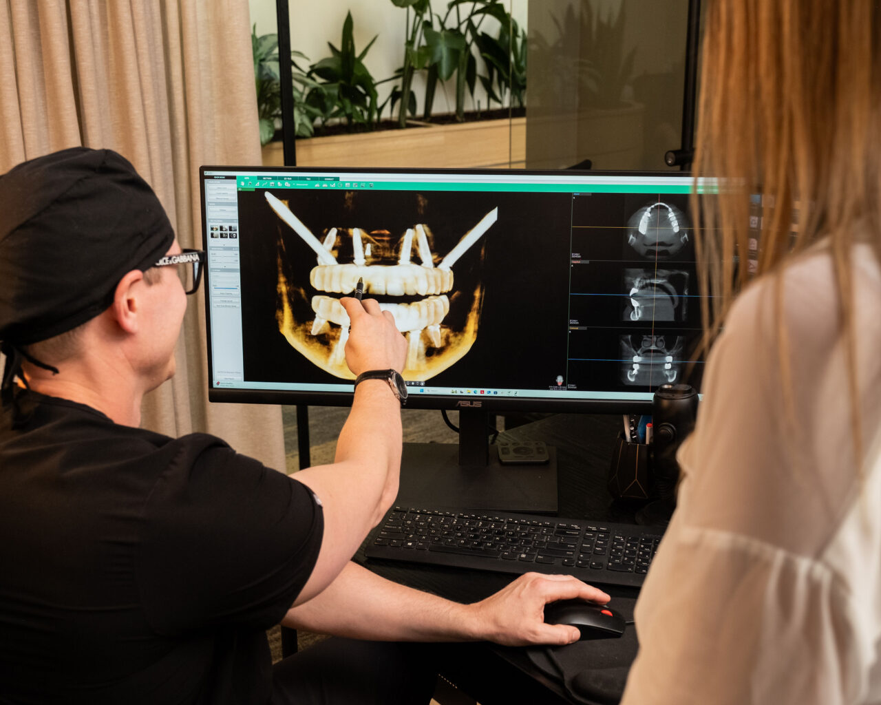 'http://A%20person%20in%20medical%20attire%20points%20at%20a%20dental%20X-ray%20on%20a%20computer%20monitor,%20showing%20a%20jaw%20with%20dental%20implants,%20while%20another%20person%20observes.%20The%20workspace%20has%20a%20keyboard,%20mouse,%20and%20indoor%20plants%20in%20the%20background.