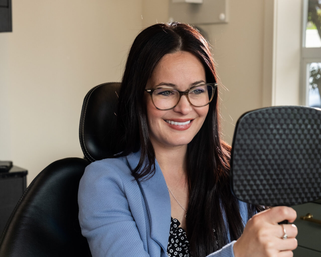 'http://A%20woman%20with%20long%20dark%20hair%20and%20glasses,%20wearing%20a%20light%20blue%20blazer,%20sits%20in%20an%20office%20chair%20smiling%20while%20looking%20at%20herself%20in%20a%20handheld%20mirror.