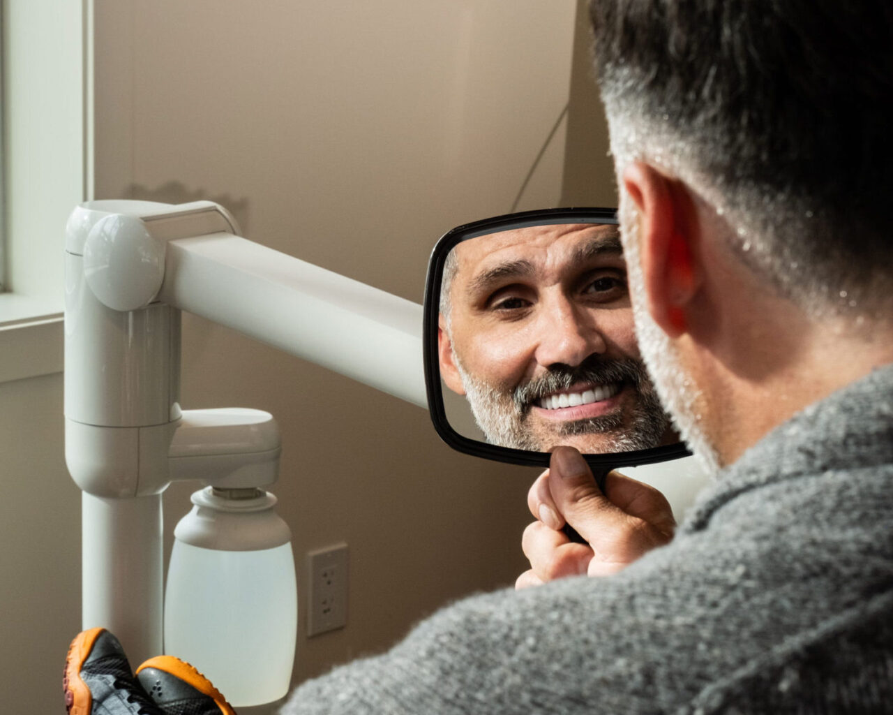 A man with salt-and-pepper hair and beard smiles as he looks at his reflection in a small handheld mirror at what appears to be a dental office.