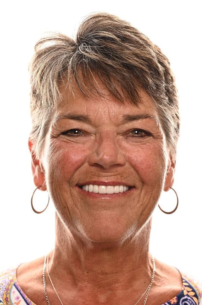 A smiling older woman with short, gray hair, wearing hoop earrings and a patterned top, against a white background.