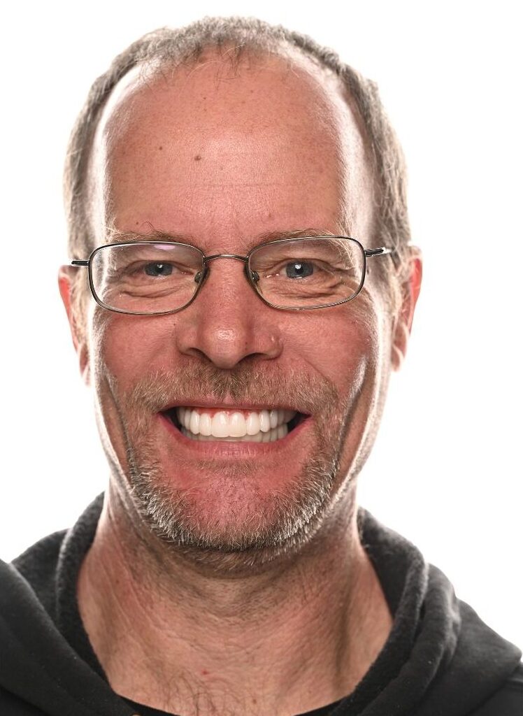 A middle-aged man with glasses, short hair, and a trimmed beard smiles broadly at the camera. He is wearing a dark hoodie and is photographed against a plain white background.