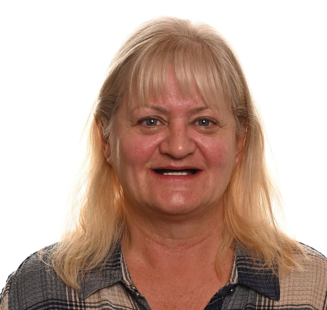 'http://A%20middle-aged%20woman%20with%20long%20blonde%20hair%20and%20bangs,%20wearing%20a%20plaid%20shirt,%20smiles%20against%20a%20plain%20white%20background.