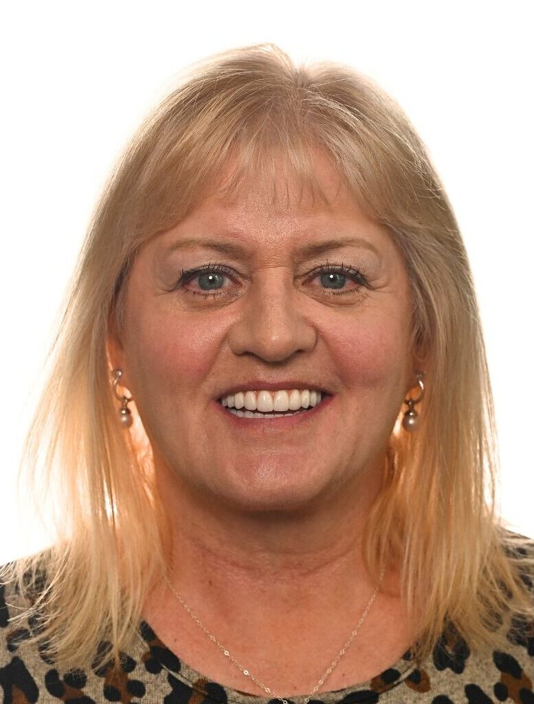 A middle-aged woman with light blonde hair, wearing a leopard print top and small earrings, smiles at the camera against a plain white background.